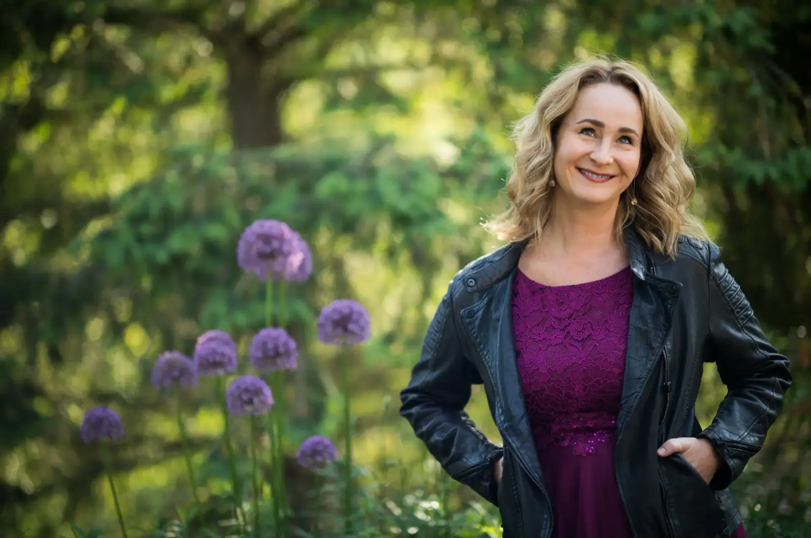 Christina Röckelein in Lederjacke und lila Kleid zwischen Allium-Blüten im Garten